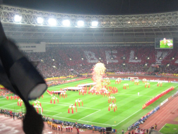 Ein Stadion mit Flutlicht, Eisengittern, Zuschauern, stehenden Personen, Werbetafeln, Anzeigetafeln, einer Sicherheitskamera und verschiedenen Fahnen und Deko.