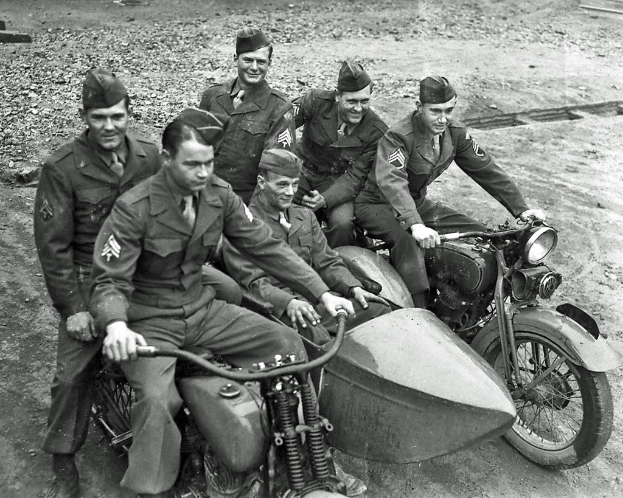 Ein Schwarz-Weiß-Foto einer Gruppe von Männern, die auf Fahrrädern sitzen und lächeln, auf einer Straße.