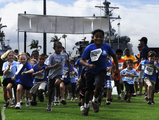 Kinder laufen bei einem Wettbewerb auf einem grünen Feld mit einem Schiff, Bäumen, Pfählen und Drähten im Hintergrund unter einem klaren blauen Himmel.