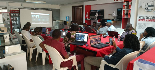 Eine Gruppe von Menschen sitzt um einen Tisch mit Laptops, Mäusen und Flaschen und nimmt an einem digitalen Marketing-Workshop teil, wobei ein Banner, ein Anzeigebildschirm und elektronische Geräte im Hintergrund zu sehen sind.