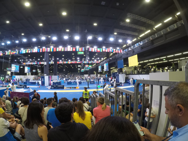 Große Menschenmenge sitzt und steht in einer Indoor-Sportarena mit Bildschirmen, Fahnen und Deckenbeleuchtung.
