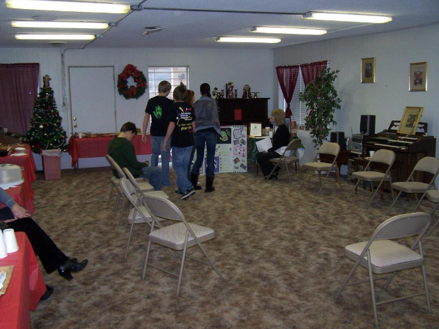 Ein Raum mit mehreren Stühlen, Tischen und Personen, einige stehen, einige sitzen, mit Tassen und Gefäßen auf den Tischen, einem Weihnachtsbaum, einem Piano auf der rechten Seite, Rahmen, Fenstern und Röhrenleuchten an der Decke und einer Wand mit einem Strauß, Vorhang, Tür und weiteren Rahmen im Hintergrund.