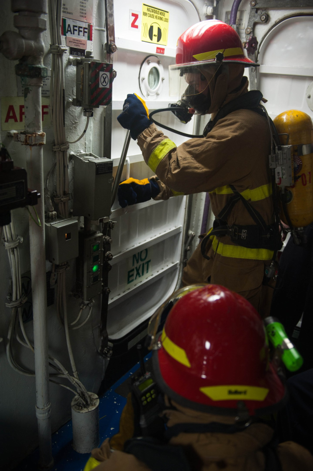 Zwei Feuerwehrleute in Schutzausrüstung inspizieren industrielle Ausrüstung mit Rohren in einer Einrichtung.