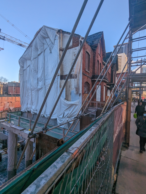 Eine Gruppe von Menschen ├╝berquert eine Br├╝cke mit Ger├╝st, mit Individuen auf einem Gehweg, einem Bauwerk im Bau, einem anderen Bauwerk, einem Kran und dem Himmel im Hintergrund.