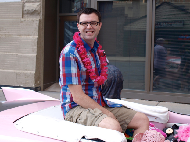 Ein Mann mit Brille und einem Lächeln sitzt in der Mitte, mit einem Fahrzeug unten, einem Gebäude und Glastüren im Hintergrund und Spiegelungen von Personen, Gebäuden und Schildern durch die Glastüren sichtbar.