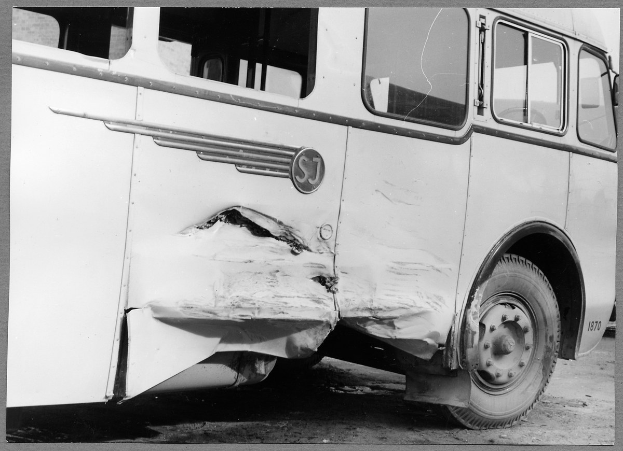 Schwarzes und weißes Foto eines verbeulten Bus mit eingedellter Front, zersplitterten Fenstern und geöffneter Fahrertür, die den Innenraum zeigt.