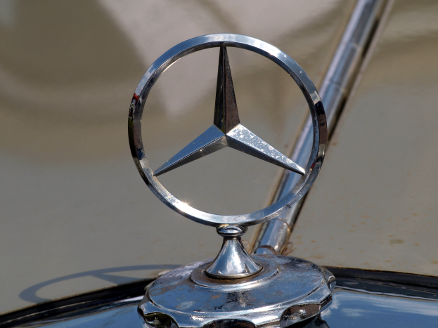Ein Close-up eines silbernen Mercedes-Logos mit schwarzem Umriss und einem weißen Stern in der Mitte, das auf der Motorhaube eines klassisch designeden Autos dargestellt ist.