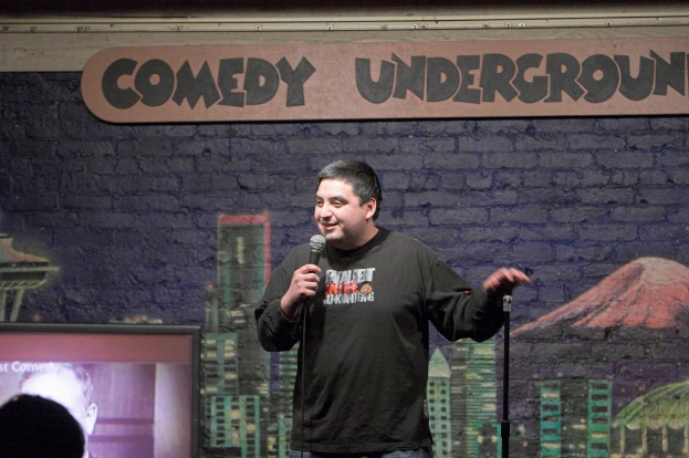 Ein Mann steht mit einem Mikrofon in der Hand und spricht, mit einem Gemälde an der Wand, einem Fernseher und Text an der Wand.