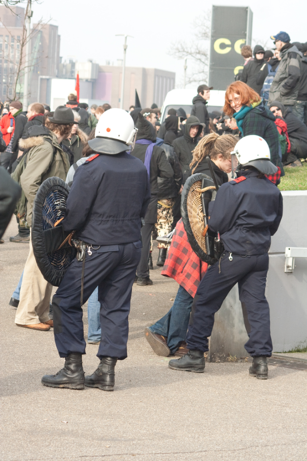 Eine Gruppe von Menschen, die auf einer Straße gehen, mit zwei Personen vorne, die wie Polizisten aussehen, Gebäuden im Hintergrund und Boden unten.
