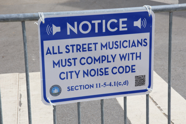 Anschlagtafel an einem Zaun mit einem Schild, auf dem "Anschlag: Alle Straßenmusiker müssen die städtische Lärmvorschrift einhalten" steht und eine Straße im Hintergrund.