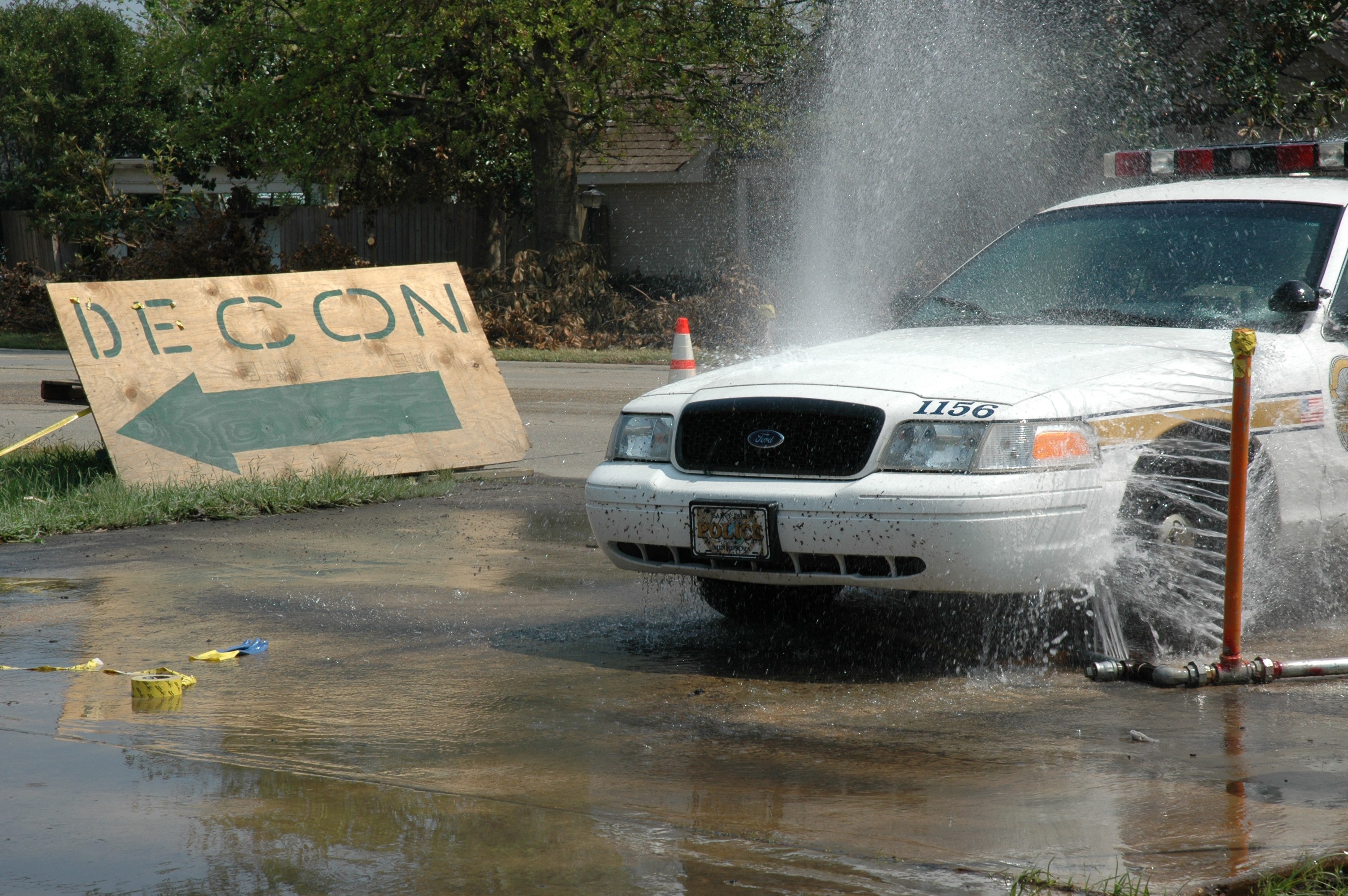 Polizeifahrzeug fährt durch eine Wasserlache mit verstreuten Gegenständen, darunter eine Tafel mit Text und ein Verkehrskegel, vor einem Hintergrund aus Bäumen, Gebäuden und einem klaren blauen Himmel.