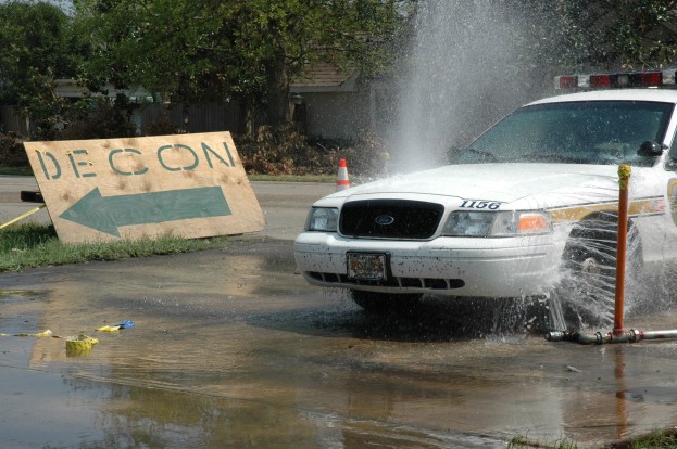 Polizeifahrzeug fährt durch eine Wasserlache mit verstreuten Gegenständen, darunter eine Tafel mit Text und ein Verkehrskegel, vor einem Hintergrund aus Bäumen, Gebäuden und einem klaren blauen Himmel.