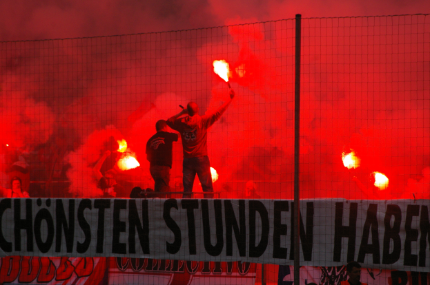 Eine Gruppe von Menschen auf einem Fussballfeld mit Fackeln, mit einem Banner, einem Zaun und einem Pfosten im Vordergrund und einem raucherfüllten Himmel im Hintergrund.