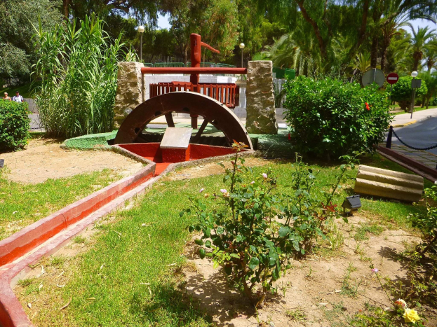 Minigolfanlage in einem Park mit Grünflächen, einer Straße, Fußgängern, Schildern, Bäumen und einem klaren blauen Himmel.