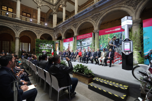 Eine Gruppe von Menschen sitzt in Stühlen und schaut auf einen Mann auf einer Bühne, der ein Mikrofon hält, mit Fahrrädern auf der rechten Seite und verschiedenen Elementen wie Plakaten, Pflanzen, Säulen, Geländern, Lampen, Fenstern und einer Wand im Hintergrund.