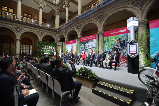 Eine Gruppe von Menschen sitzt in Stühlen und schaut auf einen Mann auf einer Bühne, der ein Mikrofon hält, mit Fahrrädern auf der rechten Seite und verschiedenen Elementen wie Plakaten, Pflanzen, Säulen, Geländern, Lampen, Fenstern und einer Wand im Hintergrund.