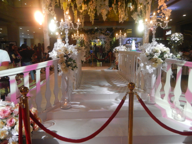 Eine Trauung in einem Einkaufszentrum mit weißem Teppich, verzierten Geländern, Blumenarrangements und Deckenbeleuchtung, bei der zahlreiche Gäste anwesend sind.