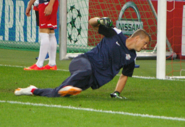 Ein Fußballspieler in Handschuhen springt, um einen Ball vor einem Tor zu fangen, mit zwei Personen, einem Netz und einer Fahne mit Text im Hintergrund.