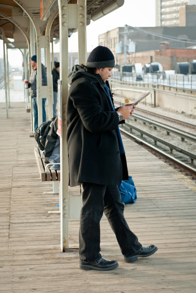 Ein Mann in einer schwarzen Jacke steht an einem Bahnhof, mit einem Eisenstab und einer Hütte im Hintergrund und einer Bahnlinie und braunen Gebäuden rechts.