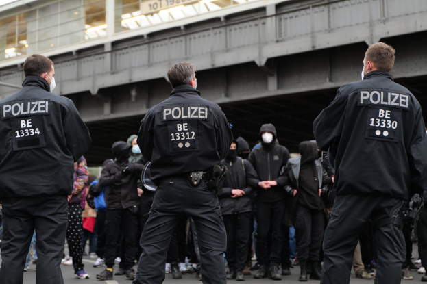 Eine Gruppe von Polizisten in Uniform steht vor einer Menge von Menschen ebenfalls in schwarzen Uniformen und Masken, mit einer Brücke und einem Gebäude im Hintergrund, während einer Demonstration in einer Stadt.