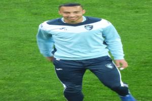 Ein Mann in einem blauen Hemd und schwarzen Hosen lächelt, während er auf einem Fußballfeld geht und Schuhe trägt.