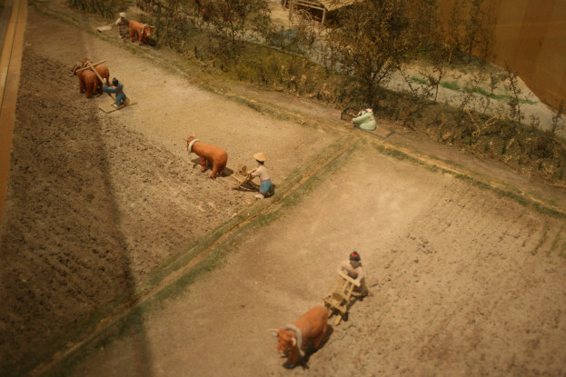 Eine Modellhofszene mit Menschen bei der Arbeit, Tieren, Pflanzen, Bäumen und einer umgebenden Wand.