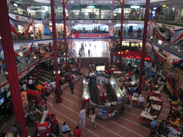 Ein belebtes Einkaufszentrum mit Säulen, Menschen, Tischen, Fliesen, Fahnen, Geschäften, einem Banner, Treppen und einem Namensschild.