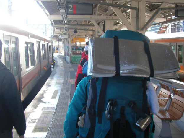 Ein Bahnhofsgebäude mit Menschen neben einem Zug, einem Stand in der Mitte, einem weiteren Zug auf der rechten Seite und einer Bank in der unteren rechten Ecke.
