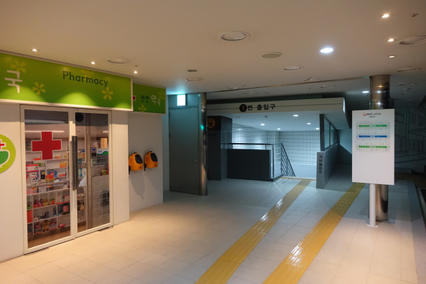 Der Eingang zu einer Apotheke mit einem 'Apotheke'-Schild auf der linken Seite, einer Tafel mit Text auf der rechten Seite und beleuchteten Lampen auf dem Dach.