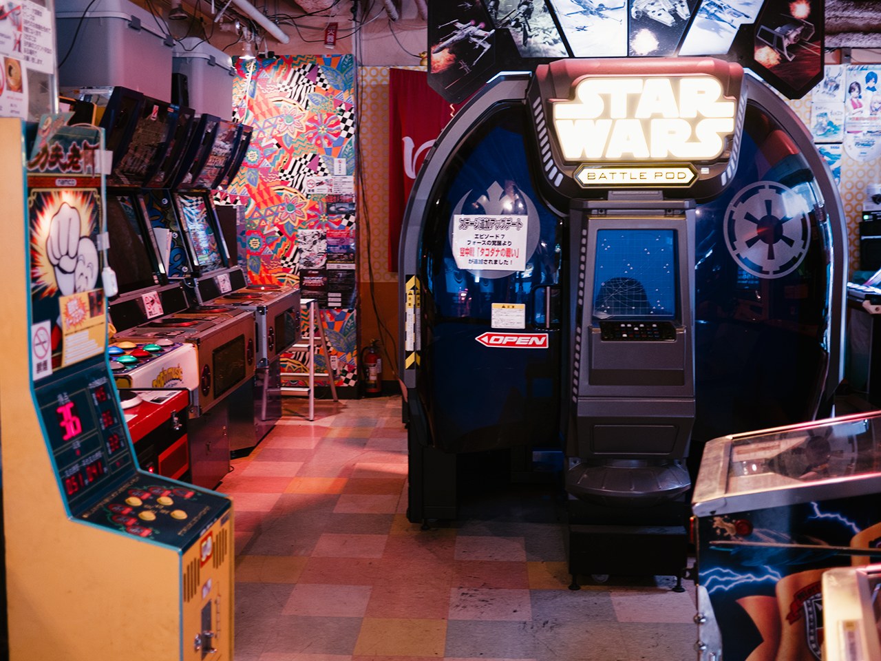 Ein hell erleuchteter Arcade-Raum mit mehreren Maschinen, darunter ein Star Wars Battle Pod, farbenfrohe Poster an den Wänden und verstreute Gegenstände.