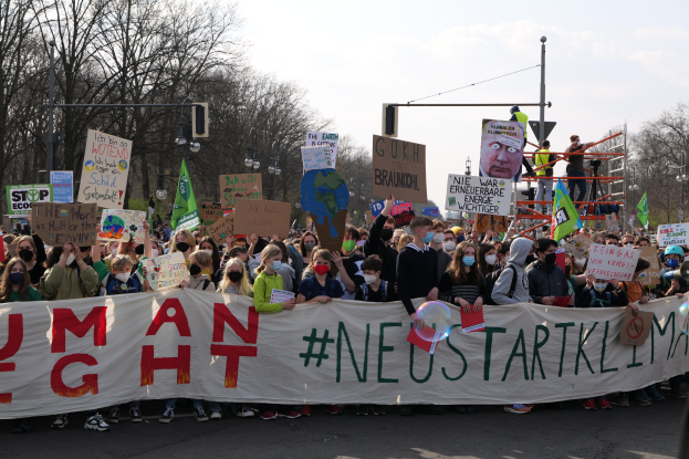 Eine große Gruppe von Menschen marschiert auf einer Straße, hält eine 'Menschenrechte'-Schleife und verschiedene Plakate, mit Bäumen, Laternen und einem klaren blauen Himmel im Hintergrund.