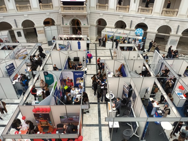 Große Gruppe von Teilnehmern auf einer internationalen Fachmesse mit Ständen, Bannern, Tischen, Stühlen und Ballons in einem Raum mit architektonischen Bögen, Säulen und einem Hintergrundbild.