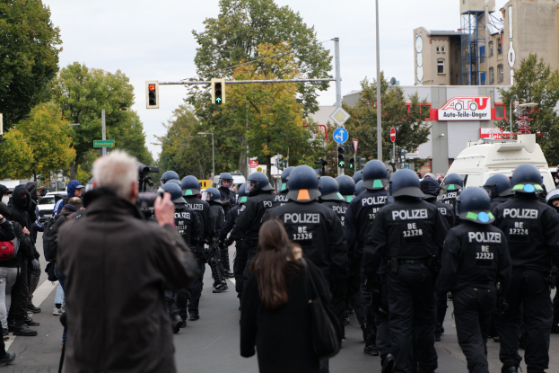 Eine Gruppe von Menschen geht eine Straße entlang mit Polizeibeamten, einige tragen Helme und haben Taschen oder Kameras bei sich, neben Fahrzeugen, Verkehrsampeln, Schildern, Bäumen, Gebäuden und einem klaren Himmel.