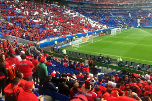 Ein volles Stadion mit Zuschauern, einige sitzen, andere stehen, mit Bannern, einem Torpfosten und Deckenleuchten, die gespannt auf den Beginn eines Fußballspiels warten.