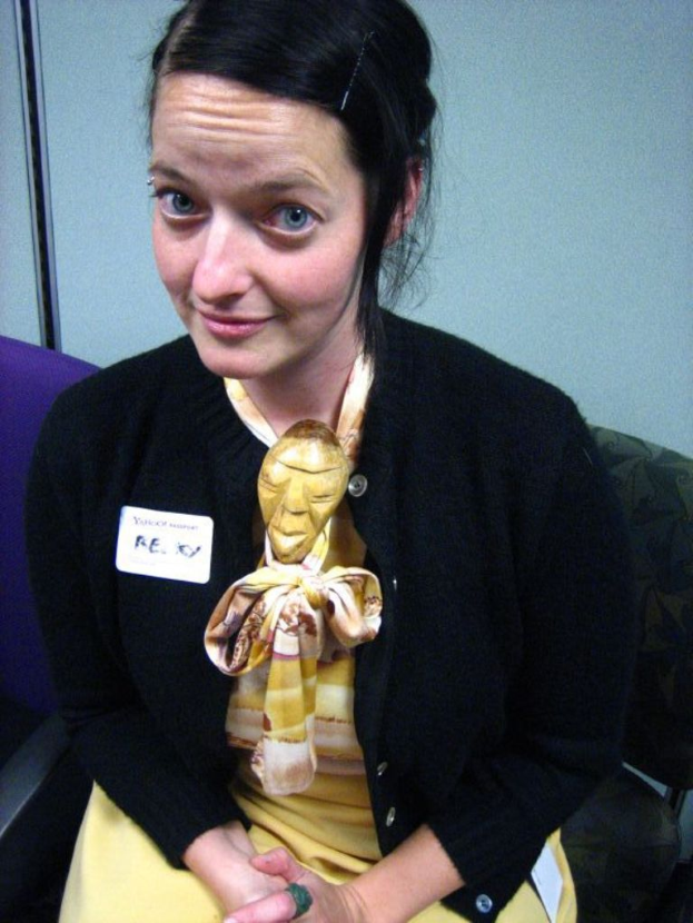 Eine Frau sitzt in einem Stuhl und trägt eine schwarze Jacke und ein gelbes Kleid, mit einem Abzeichen an ihrer Kleidung, vor einer Wand.