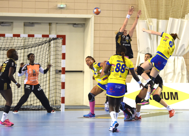 Eine Gruppe von Frauen, die Handball auf einem Platz spielt, mit einem Ball in der Luft, einem Tor mit Netz, einer Fahne mit Text, einem Vorhang und einer Wand im Hintergrund.