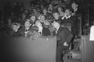 Ein Schwarz-Weiß-Foto einer Menschenmenge im Stadion mit einem Podium und einem Blumenstrauß im Vordergrund und einer Wand im Hintergrund.