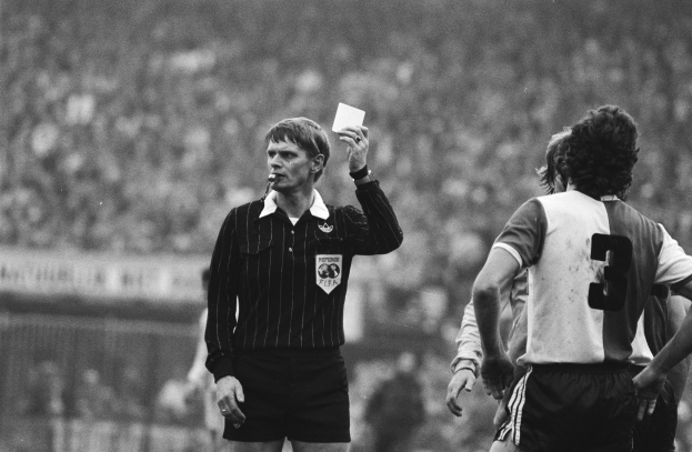 Ein Schiedsrichter in Schwarz-Weiß-Haltung hält eine rote Karte vor einer Menge von Fußballspielern, im Hintergrund ist ein Zaun zu sehen.