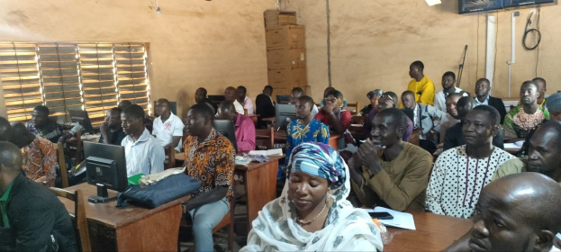 Eine Gruppe von Menschen sitzt an Tischen in einem Klassenzimmer mit Monitoren, Taschen, Büchern und Papieren und nimmt an einer Techniktrainingssitzung teil; ein Fenster ist links und Kartons und ein Fernseher sind an der Wand im Hintergrund.