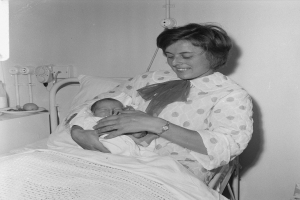 Eine Frau in einem Krankenhausbett hält ein Neugeborenes, mit einem Kissen und einer Decke in der Nähe und einem Tisch mit verschiedenen Gegenständen links daneben, mit Rohren im Hintergrund.