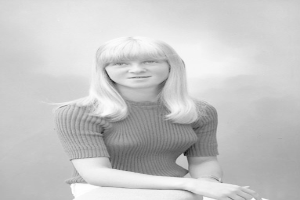 Eine Frau in einem weißen Kleid mit klassischem Bob-Haarschnitt sitzt friedlich auf einem Stuhl, die Augen geschlossen, die Hände ruhen auf ihrem Schoß, in einem Schwarz-Weiß-Foto.