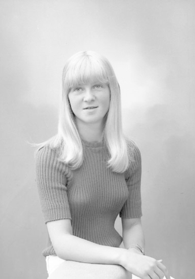 Eine Frau in einem weißen Kleid mit klassischem Bob-Haarschnitt sitzt friedlich auf einem Stuhl, die Augen geschlossen, die Hände ruhen auf ihrem Schoß, in einem Schwarz-Weiß-Foto.