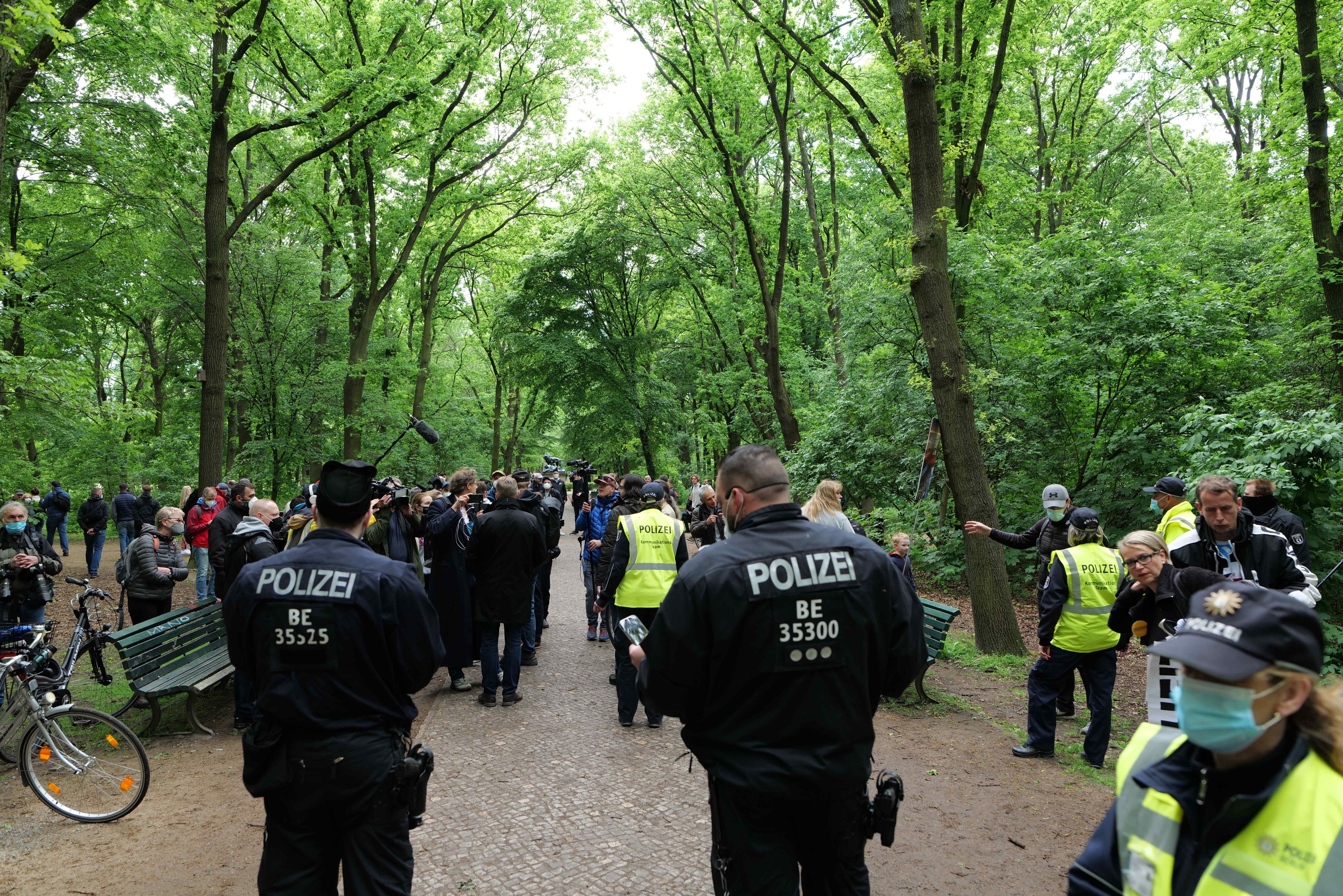 Eine Gruppe Polizisten steht vor einer Menge während einer Anti-Terror-Demonstration in Berlin, mit Fahrrädern und einer Bank im Vordergrund und Bäumen im Hintergrund.