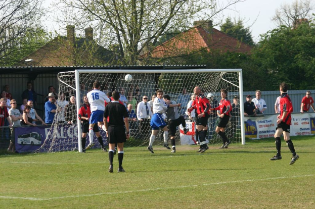 Fußballspieler auf einem Feld mit einem Tor im Hintergrund und Zuschauern dahinter, während in der Ferne Bäume und Häuser zu sehen sind.
