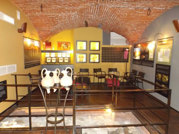 Ein Museumsraum voller Tische, Stühle, einem Geländer, Vasen, Texttafeln, Wandrahmen, Lampen und diversen Ausstellungsstücken.