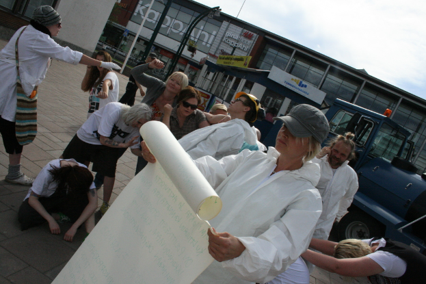 Eine Gruppe von Menschen, einige in Kostümen und Mützen, versammelt sich in der Mitte, wobei einige zu kämpfen scheinen, während eine Person ein Papier hält; im Hintergrund sind Gebäude, Pflanzen, Pfosten und Tafeln mit Text, ein Fahrzeug auf der rechten Seite, ein Weg am Boden und der Himmel oben zu sehen.