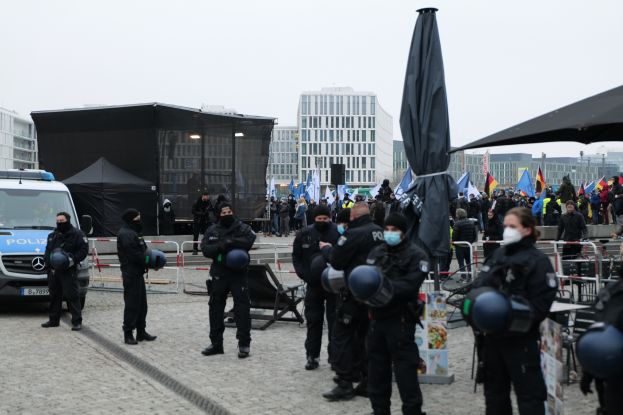 Eine Gruppe von Polizisten steht vor einer Menge von Demonstranten in Berlin, Deutschland, mit Barrikaden, Zelten und Fahnen im Vordergrund und Gebäuden im Hintergrund und einem klaren Himmel.