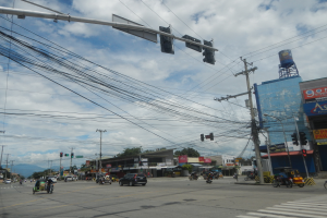 Eine belebte Stadtstraße mit zahlreichen Fahrzeugen, Strommasten mit Drähten, Gebäuden mit Namensschildern, Bäumen und einem bewölkten Himmel.
