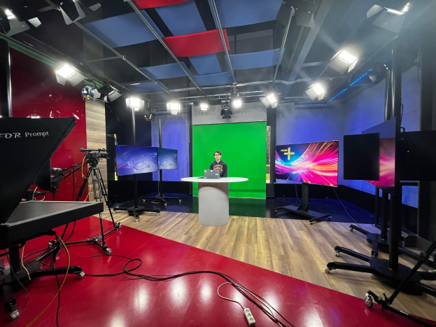 Ein Mann sitzt an einem Schreibtisch in einem TV-Studio mit einem Laptop und anderen Gegenständen, umgeben von Kameras und Equipment, mit Bildschirmen und Lampen im Hintergrund.