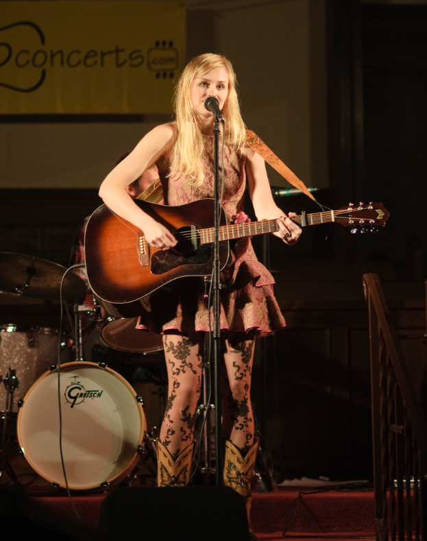 Eine Frau spielt Gitarre und singt auf der Bühne eines Konzerts, mit einer Band und einem Bildschirm im Hintergrund.